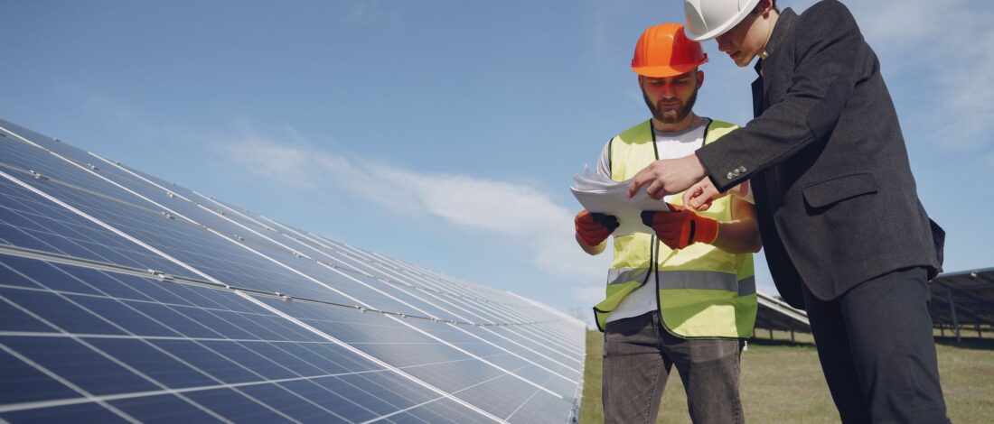 Low angle of young inspector and foreman in hardhats checking documentation against modern solar panels in field
