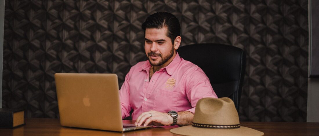Businessman working on a laptop in an office setting, wearing a pink shirt, with a hat on the desk.