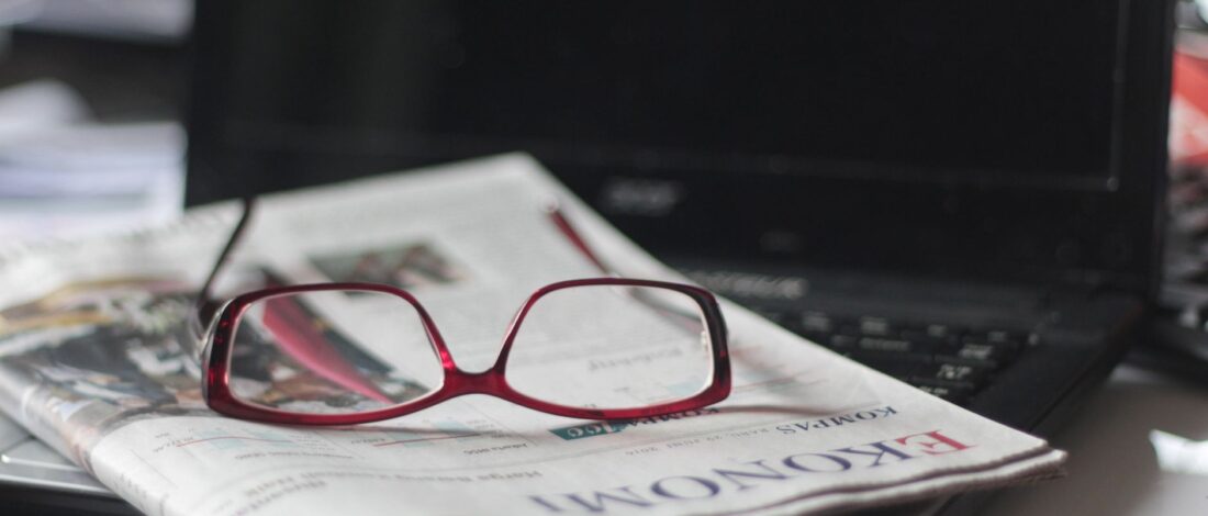 Close-up of red eyeglasses on a newspaper with a laptop in the background, suggesting reading or work.