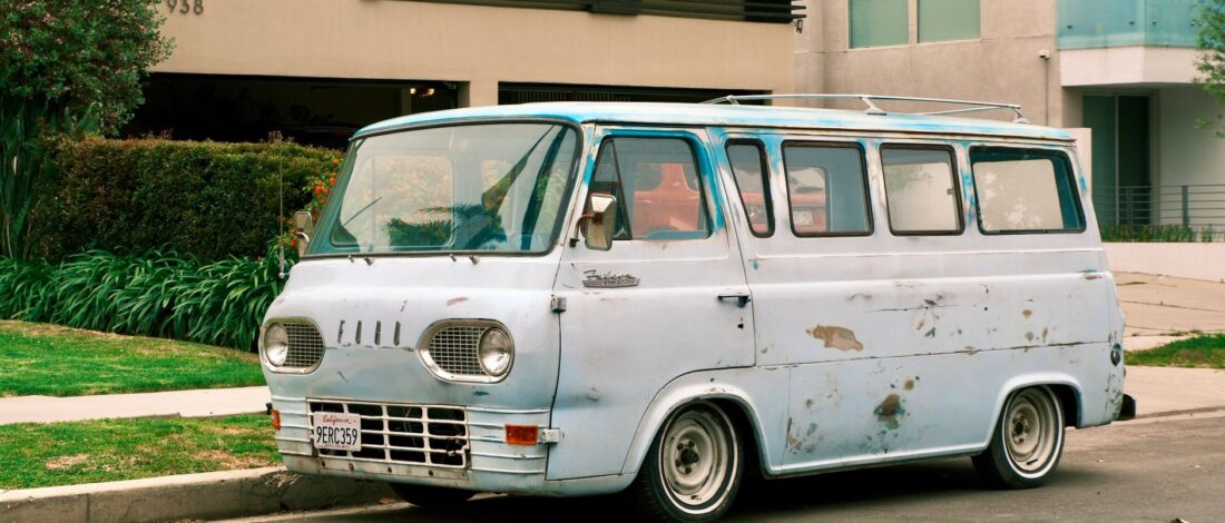 A rusty blue vintage van parked on a suburban street in front of modern buildings.