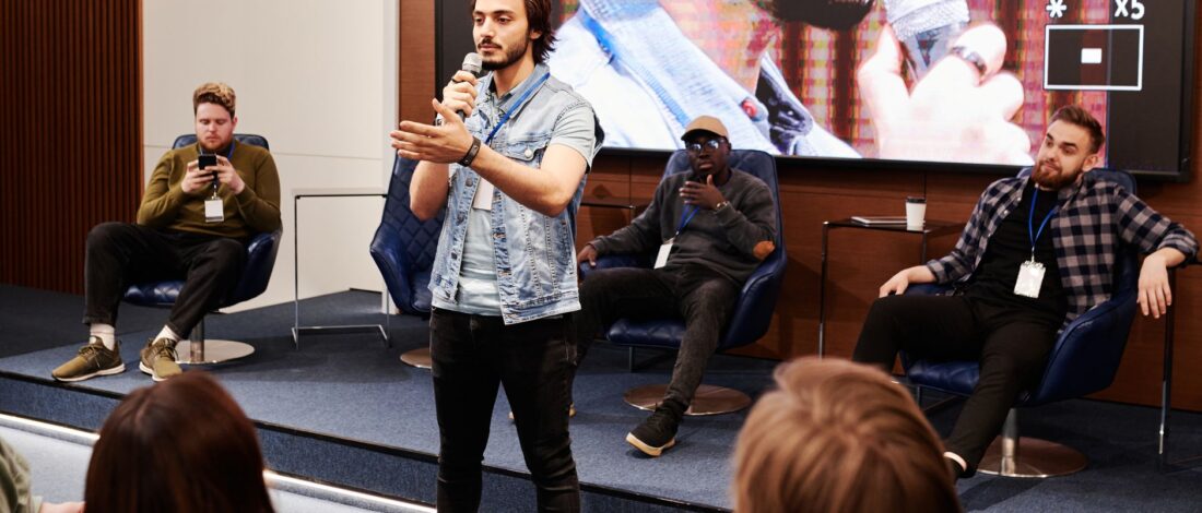 Speaker holds microphone, engaging audience at an indoor business conference with a panel in the background.