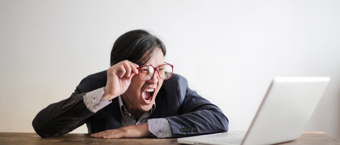 Modern Asian man in jacket and glasses looking at laptop and screaming with mouth wide opened on white background